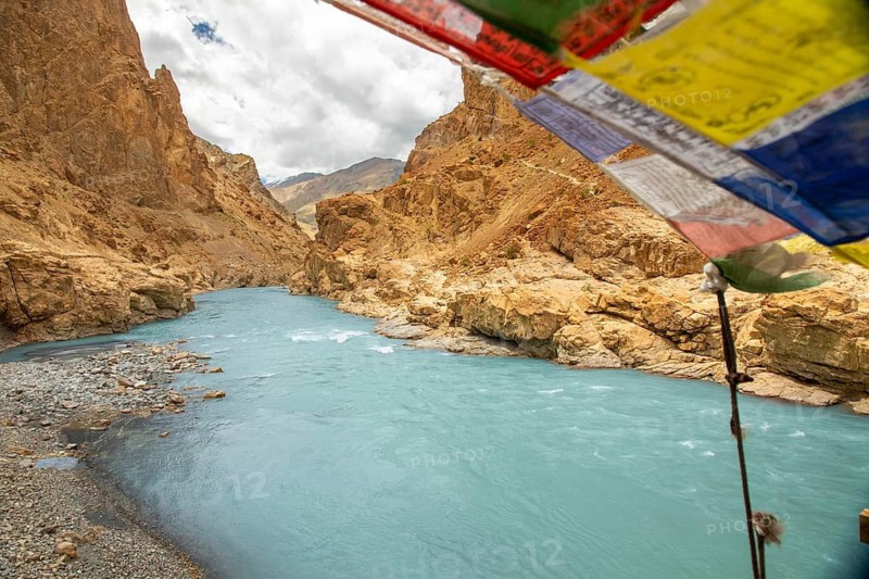 The turquoise Zanskar River winds through rugged canyons near Phugtal Gompa in Ladakh. Vibrant prayer flags flutter overhead, adding a spiritual touch to the breathtaking landscape