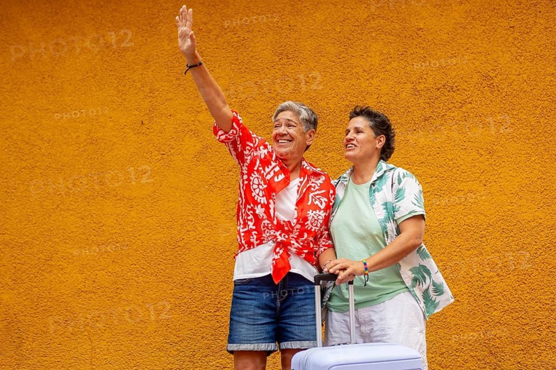 Excited lesbian couple wave and laugh ready for their summer holiday, standing beside a vivid orange wall with luggage