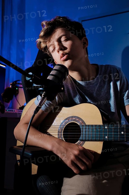 A young musician passionately sings into a microphone while playing an acoustic guitar in home studio. The blue lighting adds a creative ambiance, enhancing the musical atmosphere
