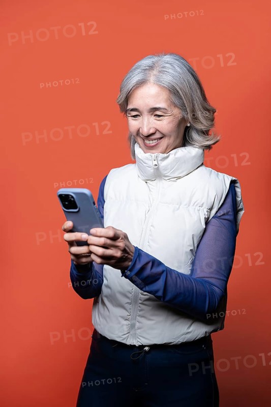 Asian Chinese senior woman enjoying technology as she smiles at her smartphone. Wearing a casual outfit, she stands against a vibrant orange background, embracing modern communication