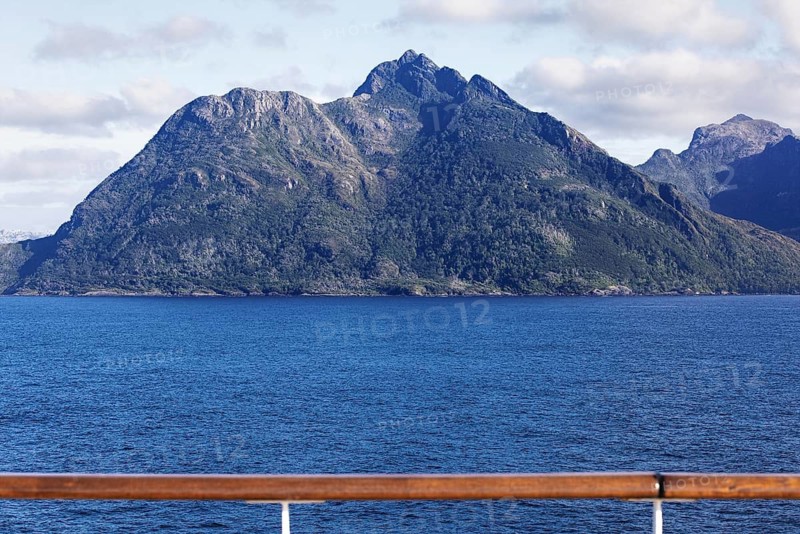 View of a distinctive, steeply rising mountain in the center of the picture taken from the deck of a cruise ship. In the foreground, deep blue sea without reflection, the ship's railing is visible at the bottom of the picture. Clear sky