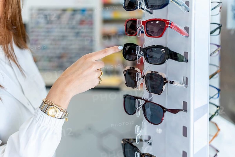 Woman's hand pointing to stylish sunglasses on a white display stand in a pharmacy, selecting eyewear for sun protection or fashion during shopping trip