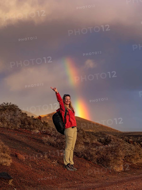 A hiker in a red jacket stands joyfully beneath a vibrant rainbow in Timanfaya National Park, Lanzarote, Canary Islands, amidst the park's unique volcanic landscape