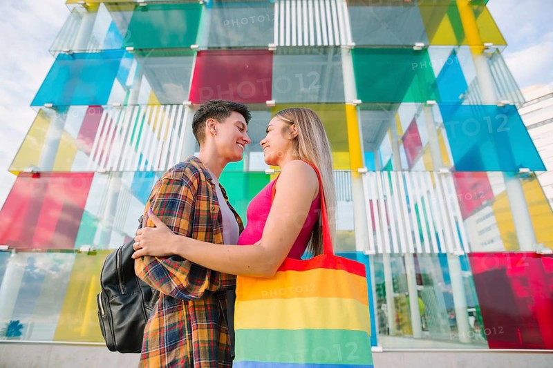 A happy lesbian couple shares a tender moment in front of a vibrant, colorful structure. They radiate love, pride, and equality, celebrating their relationship together