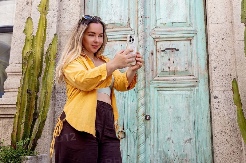 A female tourist wearing a yellow shirt takes a selfie with a smartphone in front of an old, turquoise-painted wooden door, capturing her travel experience