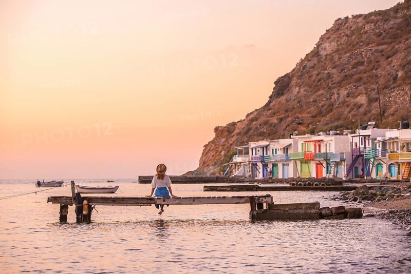 A lone woman sits on a pier in Klima, Milos Island, Greece, facing a serene sunset. Vibrant, colorful houses line the tranquil shore, evoking a sense of calm and peace