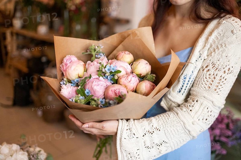 A woman in a flower shop holds a beautifully arranged bouquet of pink and white blooms wrapped in brown paper, surrounded by soft ambient lighting, creating a warm atmosphere