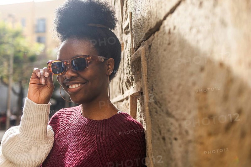 A fashionable Black woman wearing trendy sunglasses leans against a textured stone wall. Her confident smile and casual attire create a warm and sunny urban setting