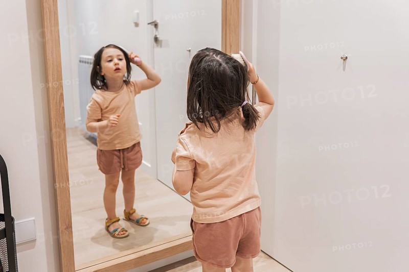Young girl stands confidently before a mirror, brushing her hair with focus and intent. She's wearing casual clothes and sandals, illustrating a moment of childhood self-care