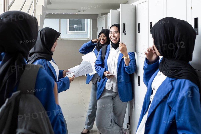 A group of smiling Asian Muslim female students in traditional attire and blue uniforms socializing in a school hallway near the lockers, expressing camaraderie and enthusiasm