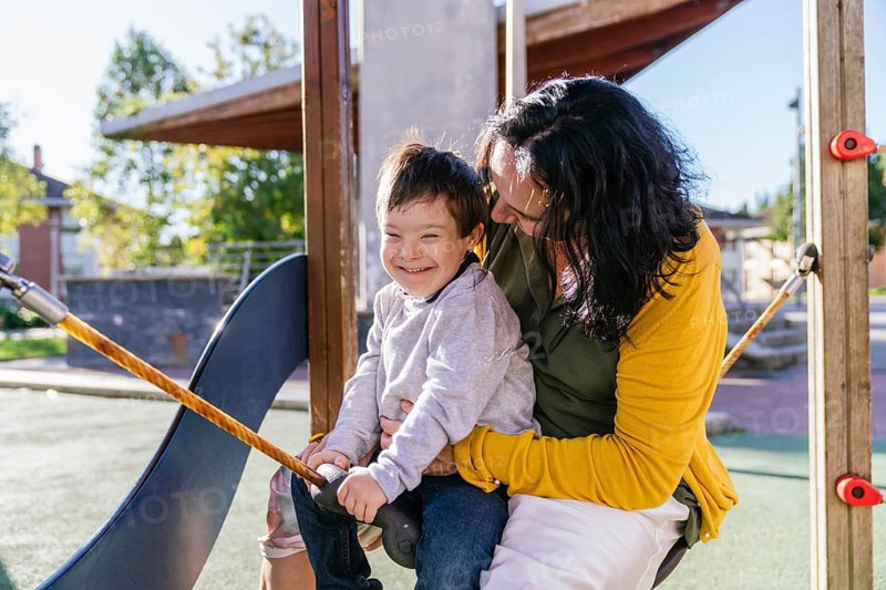A joyful mother and her smiling son with Down syndrome share a moment on a swing in a sunny playground. The scene captures warmth, love, and fun in an outdoor setting