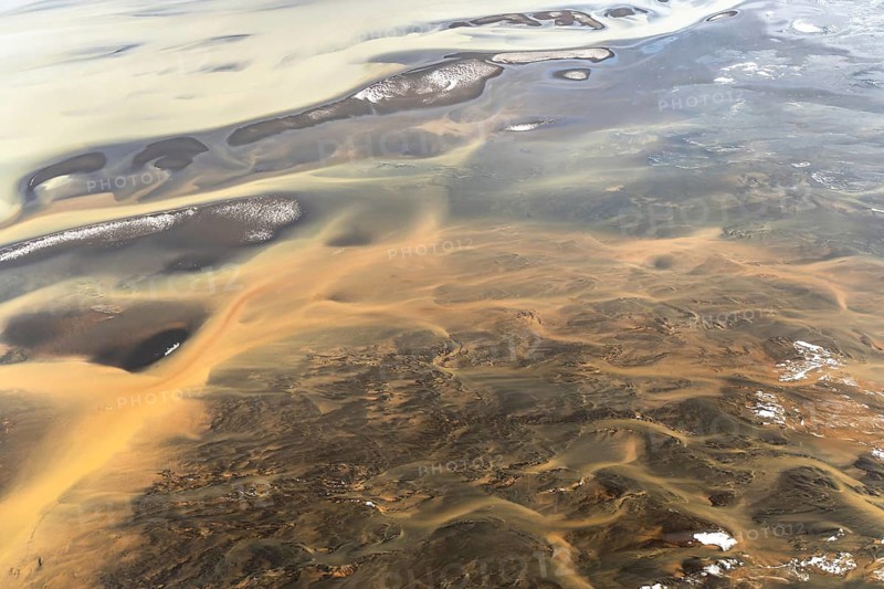 This aerial photo captures Iceland's riverside with swirling patterns and multicolored textures. The natural scene blends earthy tones with shimmering water surfaces