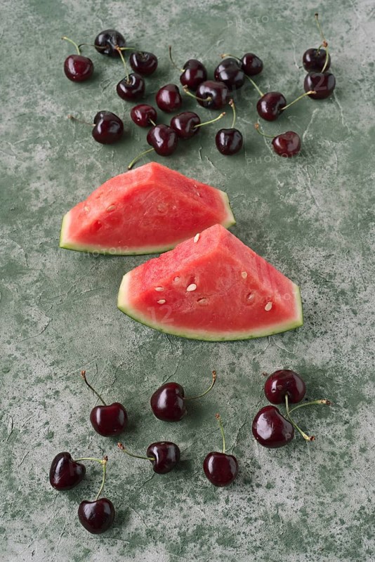 Two vibrant watermelon slices and scattered cherries on a textured green background create a refreshing, summery feel perfect for food and lifestyle visuals