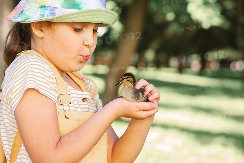 A young girl wearing a colorful hat gently holds a cute duckling in her hands, outdoors in a lush green park. She looks at the bird affectionately, enjoying a peaceful moment