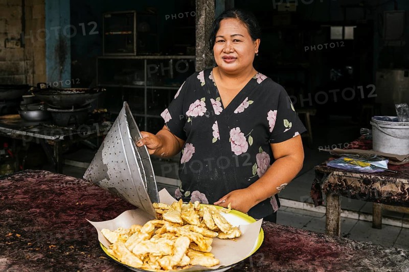 A street vendor stands behind a table displaying fresh fried snacks on a tray. She wears a floral shirt and smiles warmly, surrounded by cooking utensils in a rustic setting