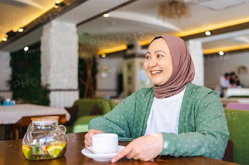 A cheerful Muslim woman wearing a hijab, seated in a cozy cafe, enjoying a cup of tea. The ambiance is inviting, with warm lighting and comfortable seating