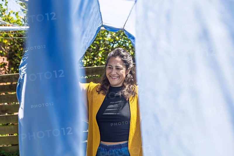 A woman stands outside, smiling as she hangs laundry on a clothesline. The sun shines brightly, highlighting the joy of simple routines in an Australian backyard