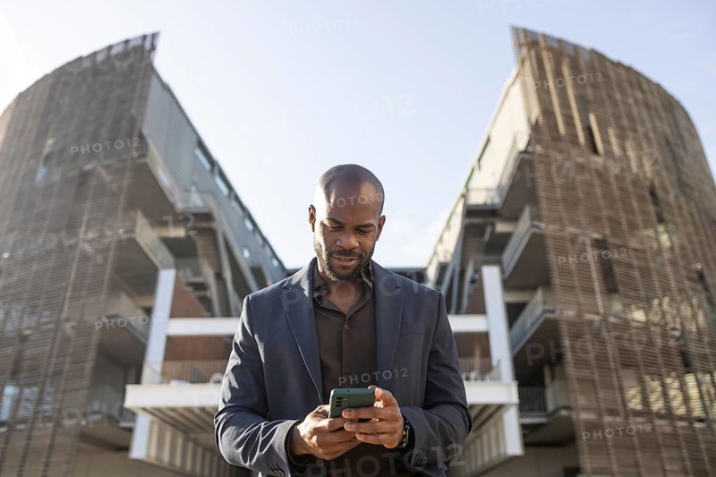 A business cuban man checks his smartphone in front of a contemporary architectural structure. He is focused and dressed in a suit, exemplifying a professional urban lifestyle