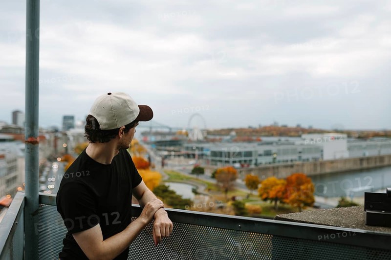 Side view of unrecognizable man in cap overlooking autumn-hued Montreal cityscape from a high vantage point. Iconic Ferris wheel featured in the distance under cloudy skies