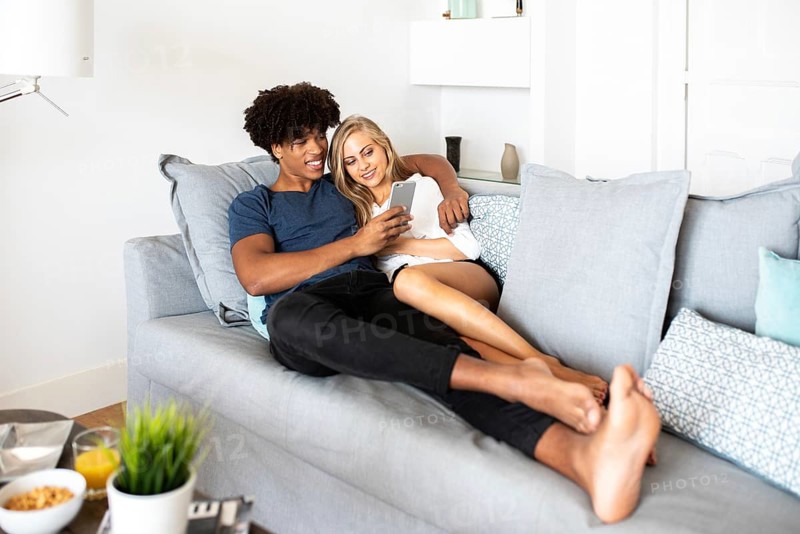 A couple cuddles on a comfortable sofa, sharing a joyful moment while looking at a smartphone. The living room setting adds to the cozy and relaxed atmosphere