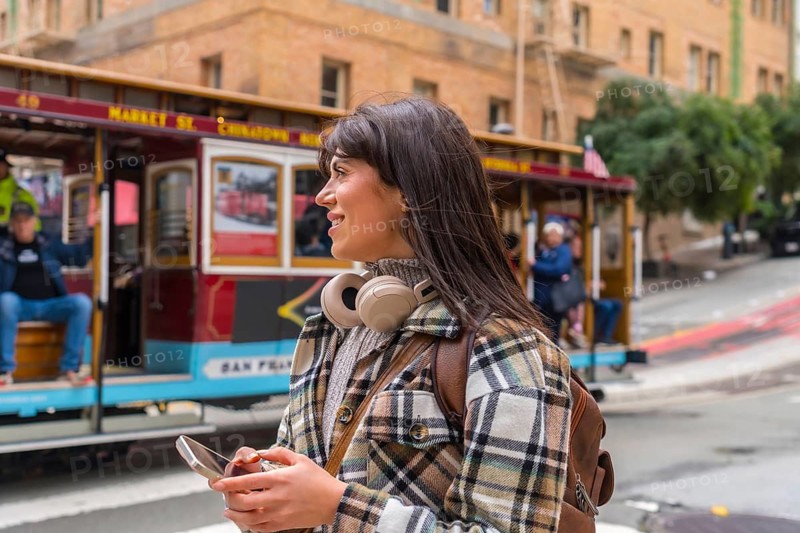 Smiling young woman exploring san francisco city, wearing headphones and backpack while holding a mobile phone, with a traditional cable car passing by in the background