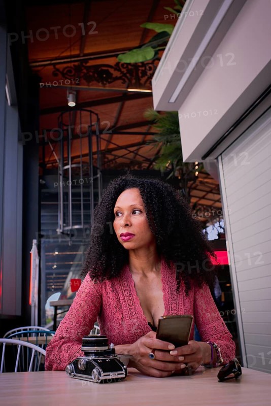 A latin woman sits at a cafe table, holding a smartphone. The ambient lighting creates a cozy atmosphere. Stylish decor and details like sunglasses and a vintage camera add elegance