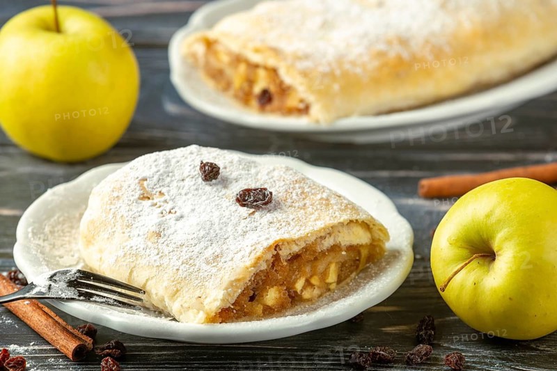 Apple strudel on plate with powdered sugar and raisins, next to yellow apples and cinnamon sticks