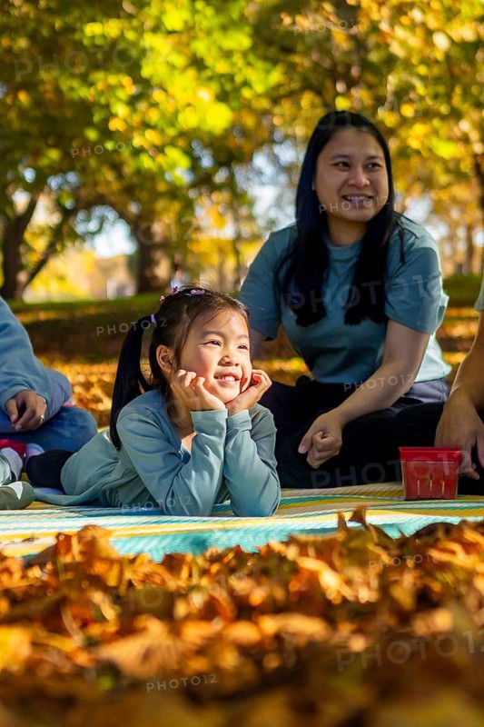 An Asian family enjoys a delightful picnic on a sunny day in a park. Surrounded by vibrant autumn leaves, they relax on a blanket, sharing laughter and joy outdoors