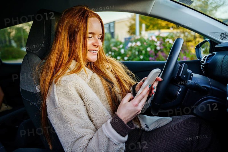 A beautiful pregnant girl with vibrant red hair sits in a car, smiling as she engages with her smartphone, surrounded by a serene daytime scene through the car window