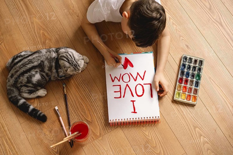 A boy lies on a wooden floor, painting a heartfelt Mother's Day card with I Love Mom. Nearby, a curious cat watches, surrounded by art supplies and bright colors