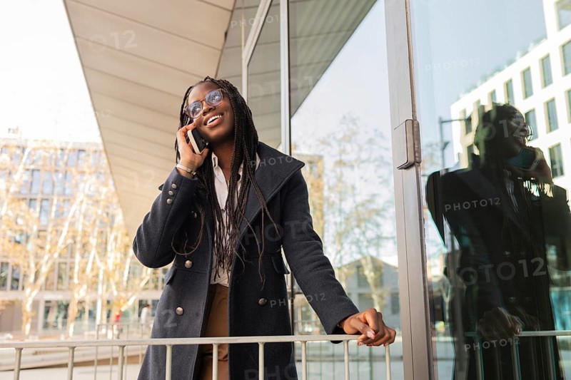 A cheerful young African American businesswoman with braids talks on her mobile phone during a break outside a modern office building