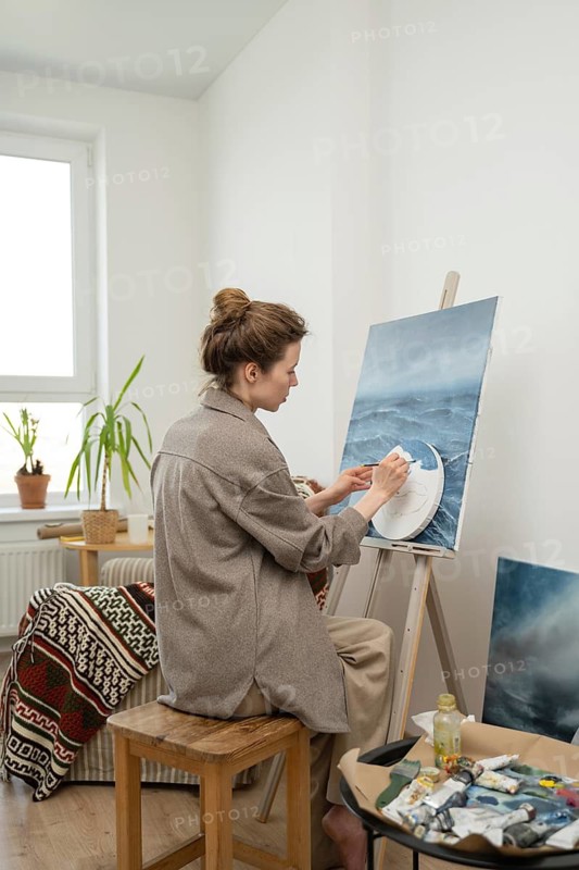 A focused artist paints on a canvas in her home studio, surrounded by natural light and cozy decor. The scene captures a moment of creativity and inspiration
