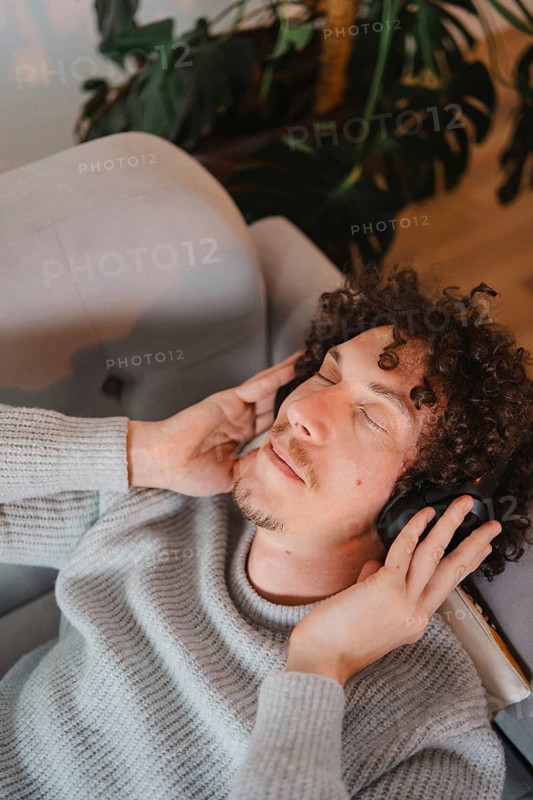 A young man with curly hair relaxes in an armchair, eyes closed, listening to music on headphones. He wears a cozy gray sweater, surrounded by lush green plants in a tranquil setting