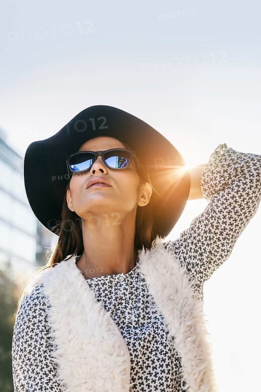Woman wearing a floppy hat, sunglasses, and textured vest. Her confident pose and bright sunlight create a stylish, carefree urban vibe. Perfect for fashion themes