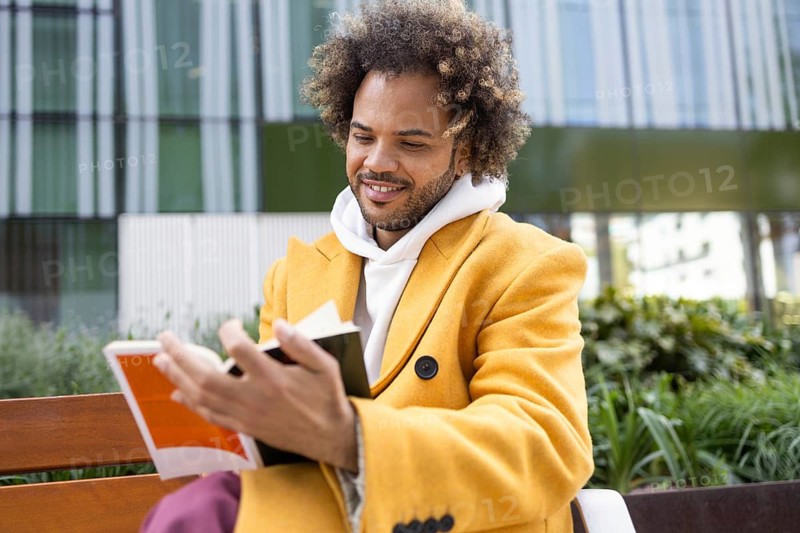 A cheerful Afro man with curly hair, wearing a bright yellow coat, enjoying a peaceful moment reading a book on a bench in an urban outdoor setting