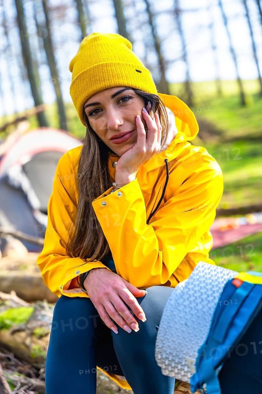 Young woman wearing yellow raincoat and beanie resting near her tent in forest during camping trip