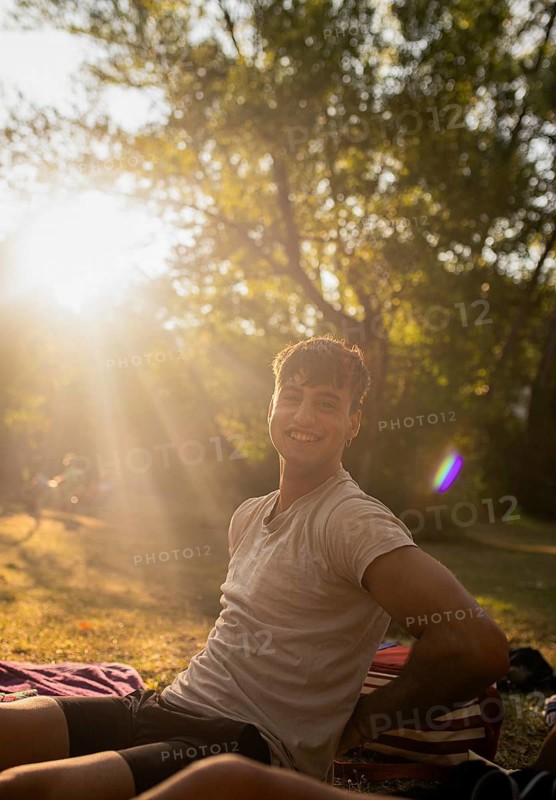 A cheerful young man enjoys the warm ambiance by the lake, sitting on a blanket with sunlight filtering through trees behind him, creating a perfect summer scene duting sunset