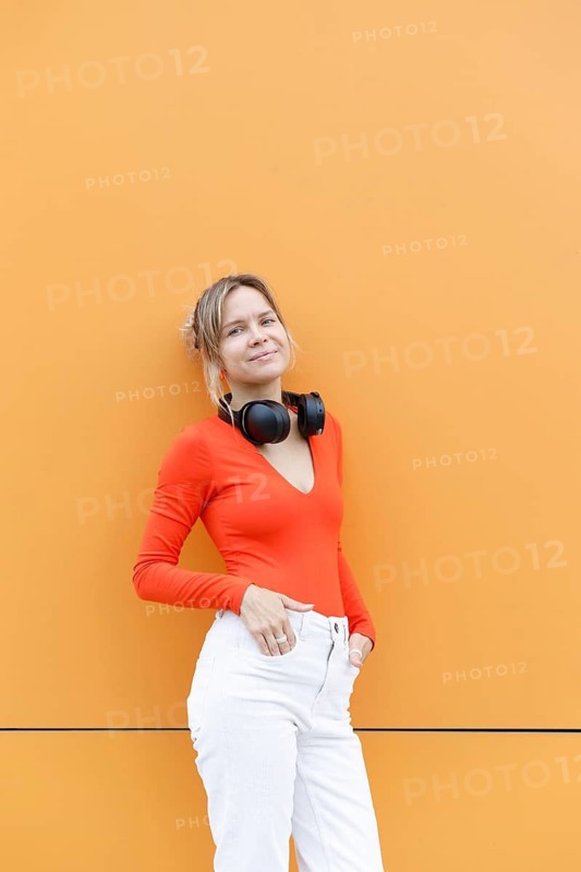 A woman in a red top and white pants leans against an orange wall in Warsaw, with headphones around her neck The vibrant background enhances her modern, stylish look