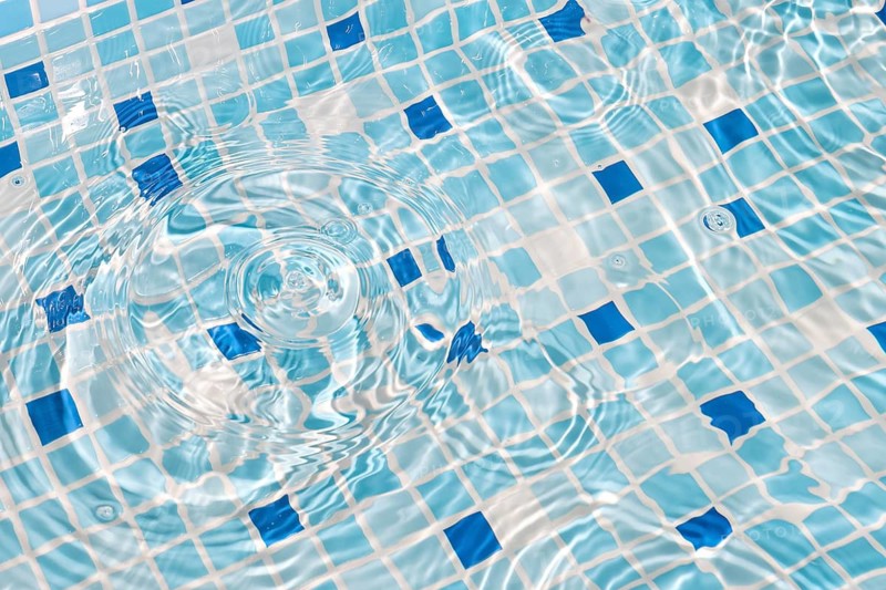 A close-up image of mosaic tiles under clear, rippling water, creating a serene and refreshing summer vibe. The blue and white tiles enhance the calming effect, with sunlight reflecting beautifully