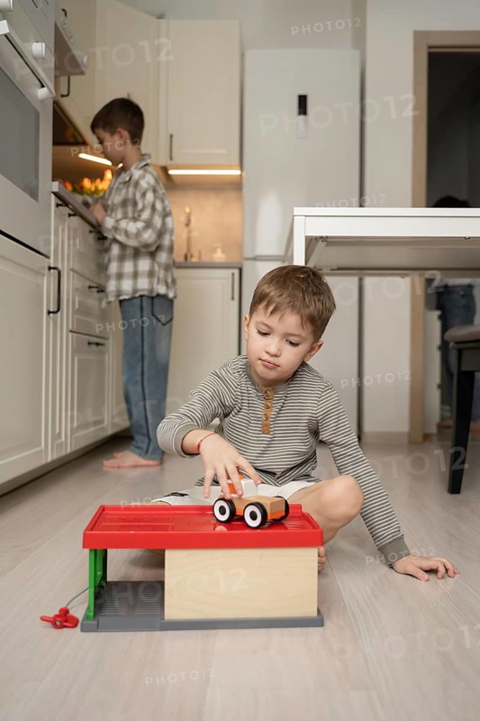 A young boy plays with a toy car on the kitchen floor, while his brother is engaged at the counter. The bright, cozy kitchen creates a warm, playful atmosphere