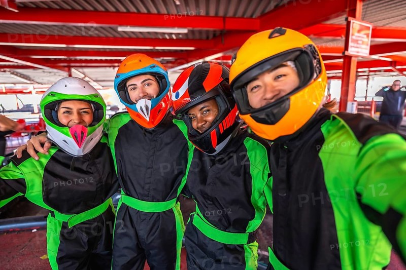 Diverse friends wearing helmets and racing suits, smiling and taking a selfie together at an indoor karting track, preparing for a fun and exciting race