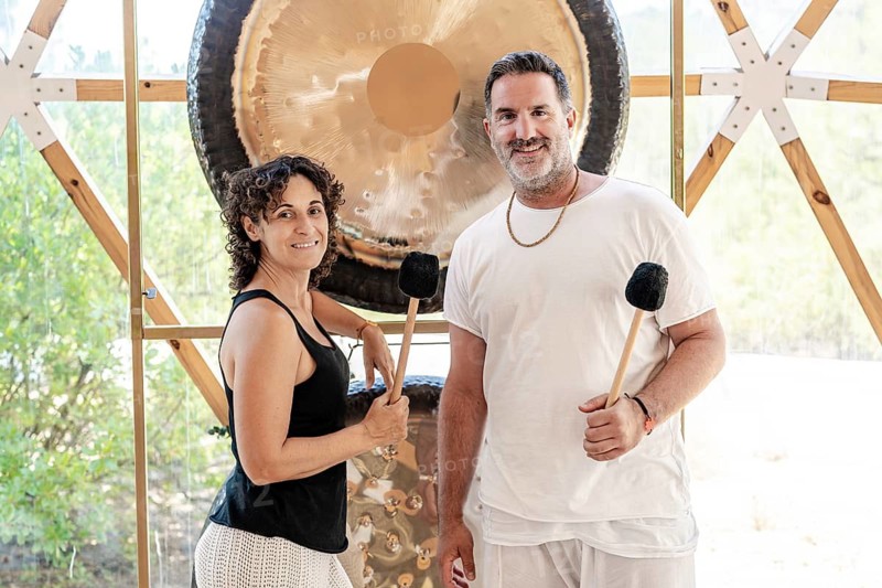 A man and woman, both sound therapists, standing looking at camera while engaged in a therapeutic gong bath session. They hold mallets and stand in front of large gongs in a serene setting