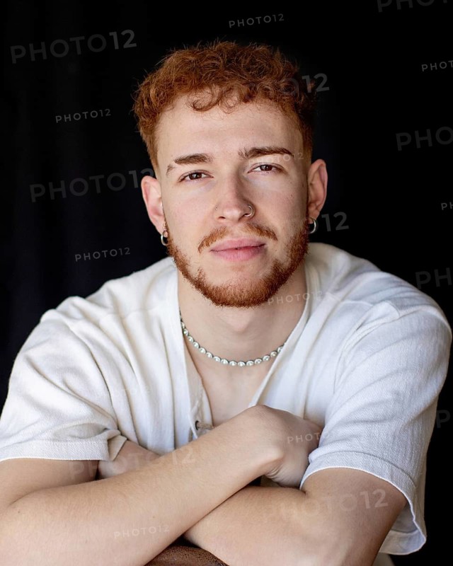 A young man with red hair and a confident expression poses indoors. He wears a white shirt, hoop earrings, and a necklace, gazing directly at the camera with arms crossed