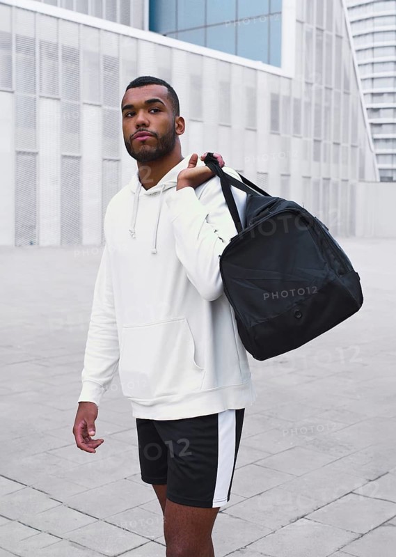 A confident black male athlete stands in an urban setting, wearing a white hoodie and shorts. He carries a black gym bag over his shoulder, exuding strength and style
