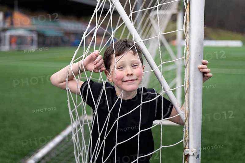 A joyful young boy playfully stands behind a soccer goal net on a lush green sports field. His cheerful expression and casual pose capture the essence of childhood fun