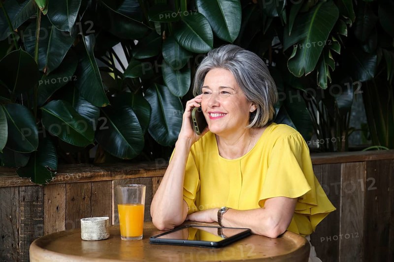 A mature woman with gray hair smiles warmly while talking on her phone at a cafe. She is wearing a bright yellow blouse, with orange juice and a tablet on the wooden table
