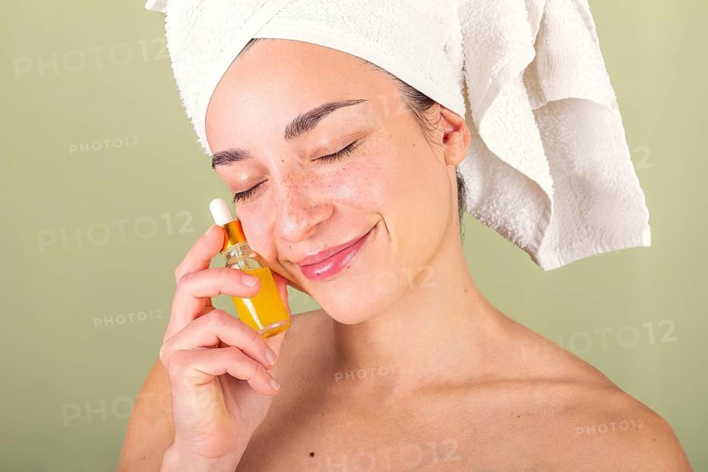 A woman with a towel wrapped around her head gently holds a bottle of facial oil against her cheek, enjoying her skincare routine. Her eyes are closed in relaxation