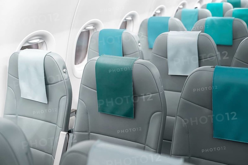 The image showcases the interior of an airplane cabin, featuring rows of modern grey seats with teal accents. Sunlight filters through the windows, highlighting the spacious and clean design