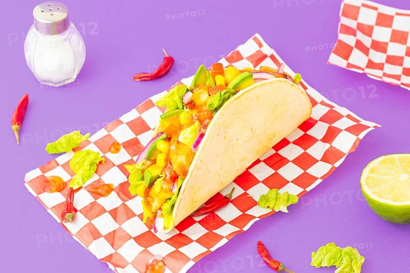 From above chicken taco filled with lettuce, corn, avocado, and red onion, styled on a red checkered paper over a purple background. Studio lighting enhances the vivid colors and textures