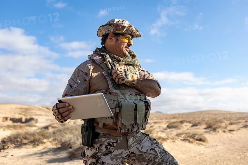 A military professional stands in a desert environment, equipped with tactical gear, holding a tablet. The image highlights the integration of technology in remote operations